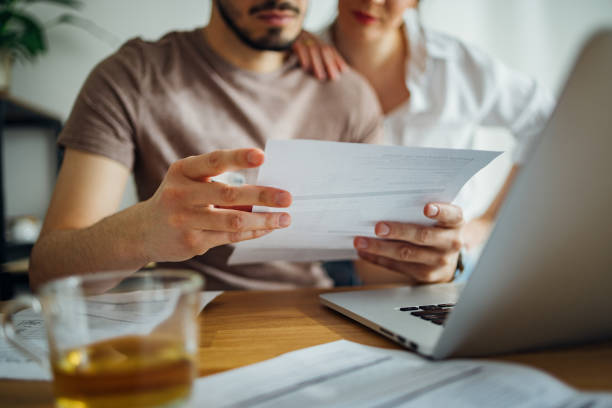 Close up photo of male hands holding bill and pen and female hands hugging him while they doing home finances together online on a laptop computer and drinking cup of tea in the kitchen.