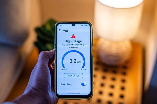 Hand holding smartphone displaying high energy usage alert from smart plug, indicating excessive electricity consumption compared to usual levels, prompting homeowner to take action