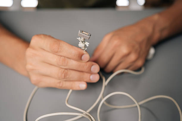 Skilled technician repairing frayed white charging cable, fingers precisely knotting damaged cord on minimalist gray workspace, showcasing technical expertise. Concept of computer hardware