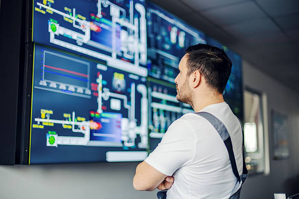 Male engineer stands with arms crossed in a modern control room, monitoring large screens showing data visualizations, graphs and schematics of industrial automation systems