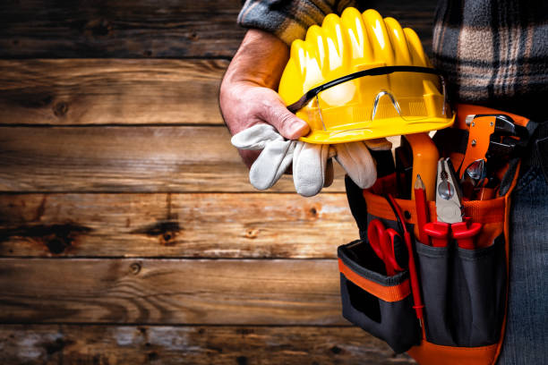 Electrician worker on vintage wooden background; holds helmet, gloves and goggles in hand. Construction industry, electrical system.
