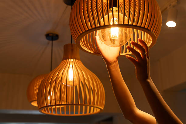 Creative hands adjusting glowing bulb in wooden pendant lamp, cozy modern interior lighting design, reflecting stylish and inspired living with natural materials, warmth, and artistic atmosphere
